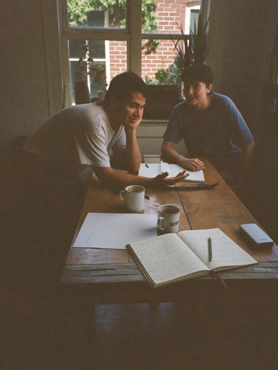Two people in conversation across a worktable covered in notebooks